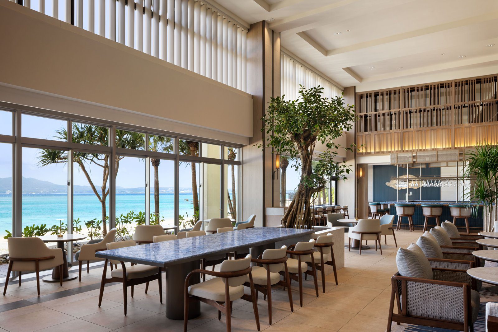 沖繩萬怡度假酒店 THE LOUNGE 挑高空間的長桌座位與吧台，落地窗外是海灘