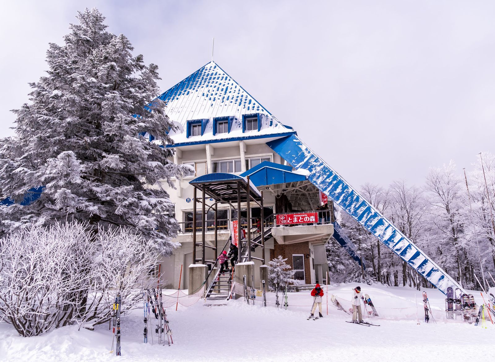 藏王纜車山麓站外觀，藍色尖頂建築與被雪包覆的針葉樹，站前月台立著滑雪板
